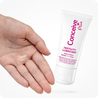 The image of Conceive Plus try me size fertility lubricant 30ml tube and a hand with clear white lube on its palm showing required dosage on a white background. The tube is white with pink branding and shows information about quantity of lube, ingredients, and made in France text.